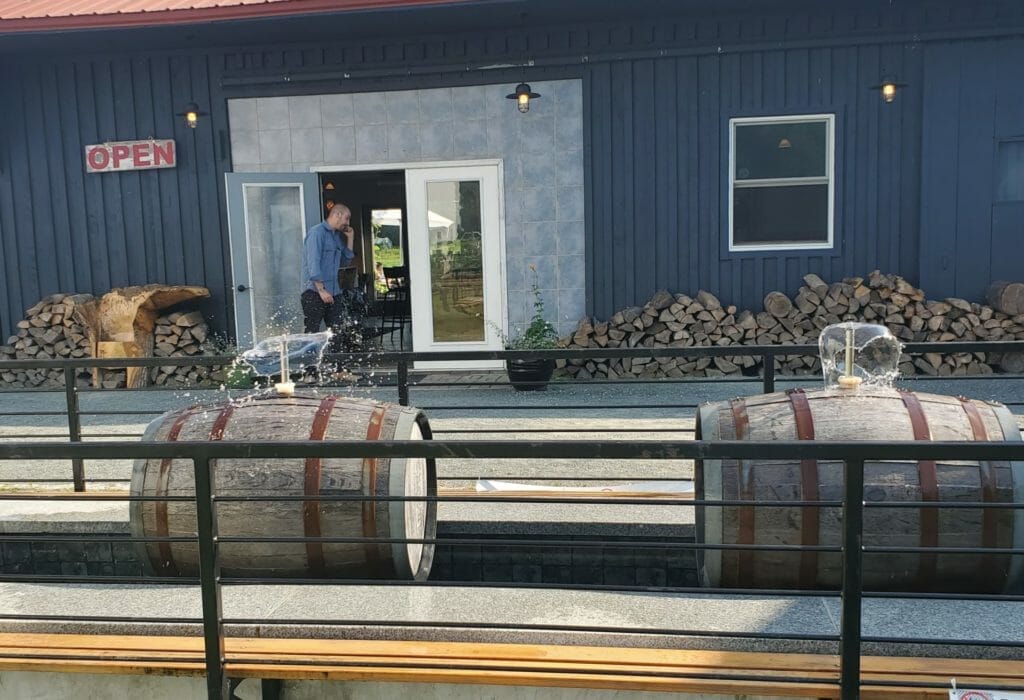 A man stands outside a building with an "OPEN" sign, next to two large barrels with water fountains on top. A stack of firewood is seen along the wall in the background.