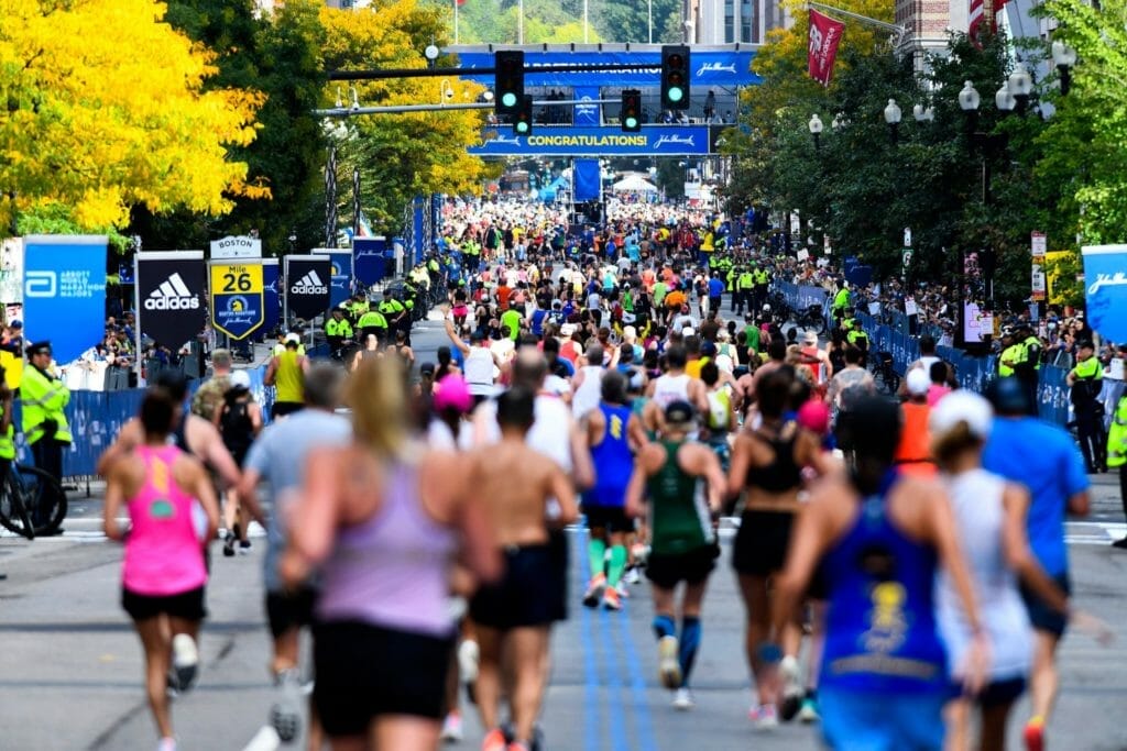 Runners approach the finish line during a marathon, surrounded by spectators and officials. A large "Congratulations" banner is displayed above the street, and the scene is bustling with activity.