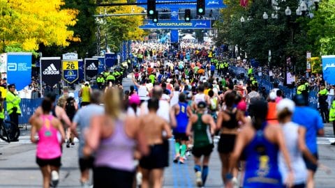 Runners approach the finish line during a marathon, surrounded by spectators and officials. A large "Congratulations" banner is displayed above the street, and the scene is bustling with activity.