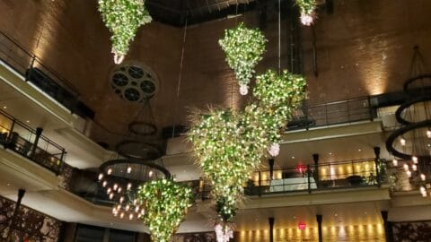 A large atrium with multiple hanging light fixtures shaped like upside-down cones, adorned with greenery and lights. The space has brick walls and multiple stories with balconies.