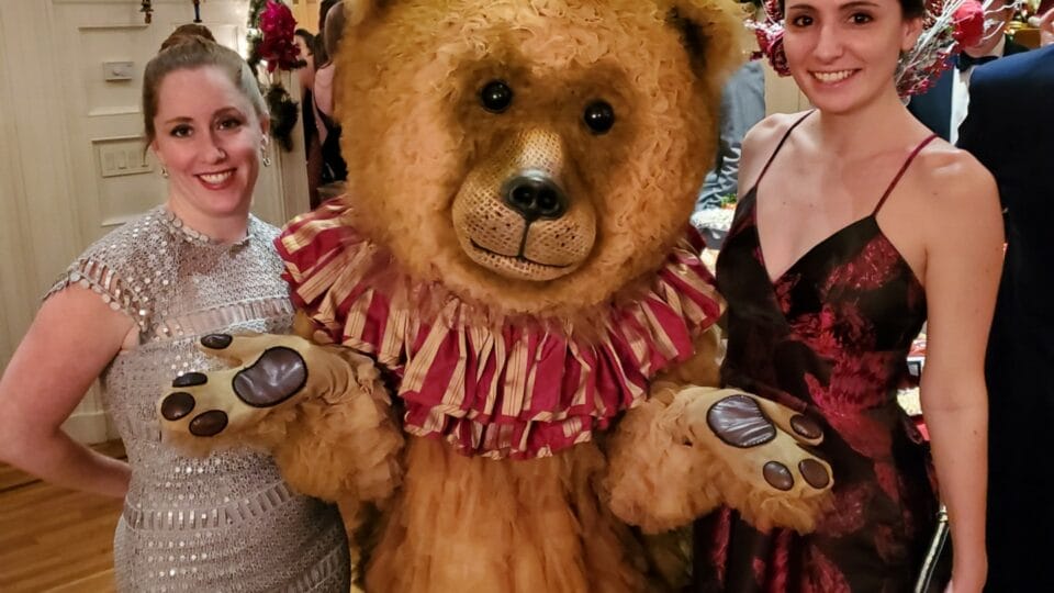 Two women pose with a large teddy bear mascot in a festive indoor setting decorated with greenery and string lights. The women are dressed in elegant evening gowns.