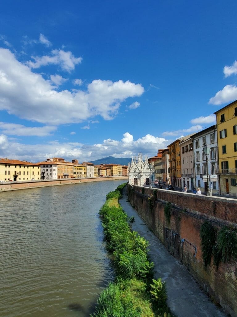 A view of the Arno River in Pisa, Italy, with colorful buildings lined along the riverbank and a small church in the distance under a blue sky with scattered clouds.