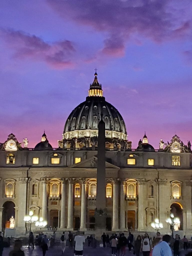 The image shows St. Peter's Basilica at dusk, with its illuminated dome and facade against a backdrop of vibrant purple and pink sky. People are visible in the foreground.