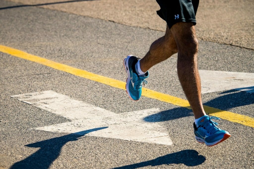 Close-up of a person jogging on a road, wearing blue athletic shoes and black shorts, with a yellow line and a white arrow visible on the asphalt.