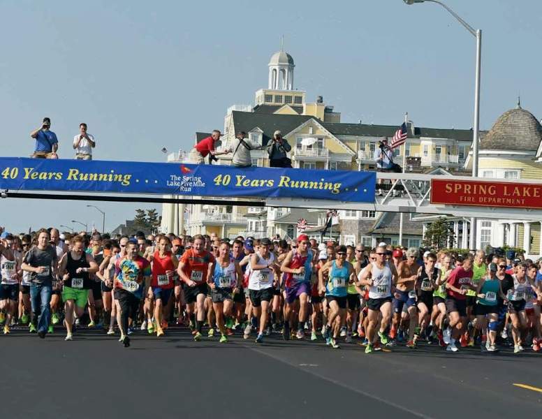 A large group of runners participate in a road race, passing under a blue banner that reads "40 Years Running," in front of a building labeled "Spring Lake Fire Department.