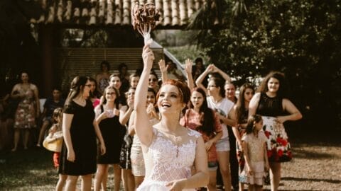 A bride in a white dress is tossing a flower bouquet into the air during one of those memorable Cape Cod wedding events, as a group of women and girls joyfully gather behind her, preparing to catch it. The scene is outdoors with lush greenery in the background.