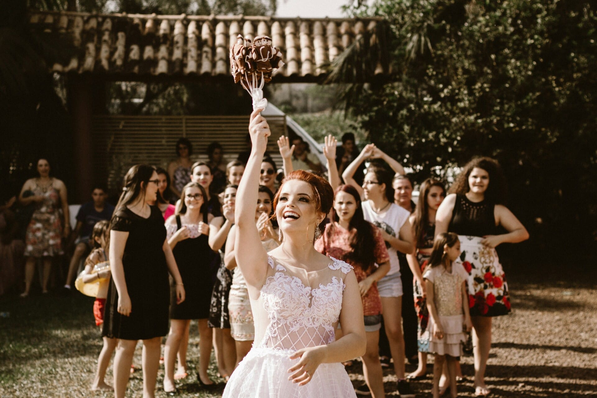 A bride in a white dress is tossing a flower bouquet into the air during one of those memorable Cape Cod wedding events, as a group of women and girls joyfully gather behind her, preparing to catch it. The scene is outdoors with lush greenery in the background.