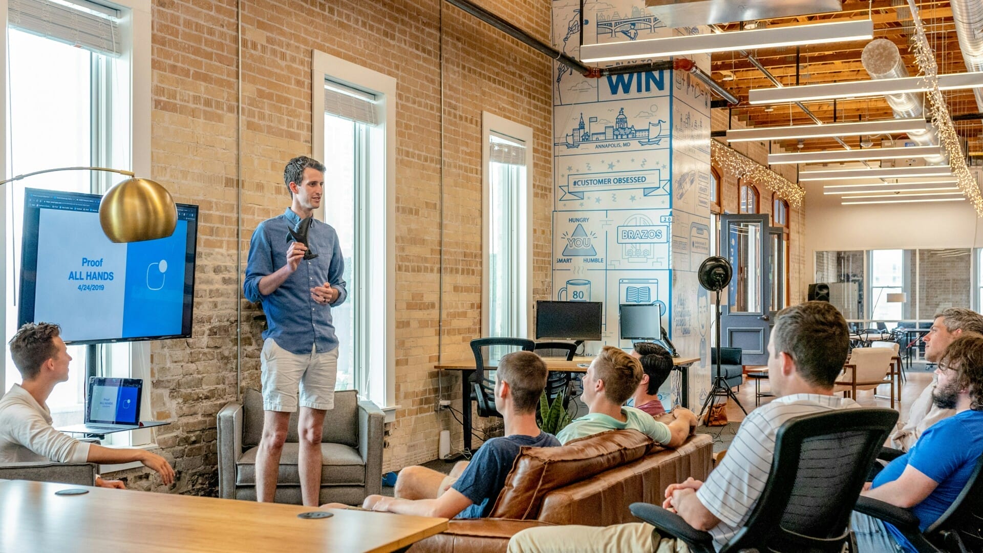 A man stands and speaks in front of a seated group in a modern office space, with a presentation displayed on a screen.