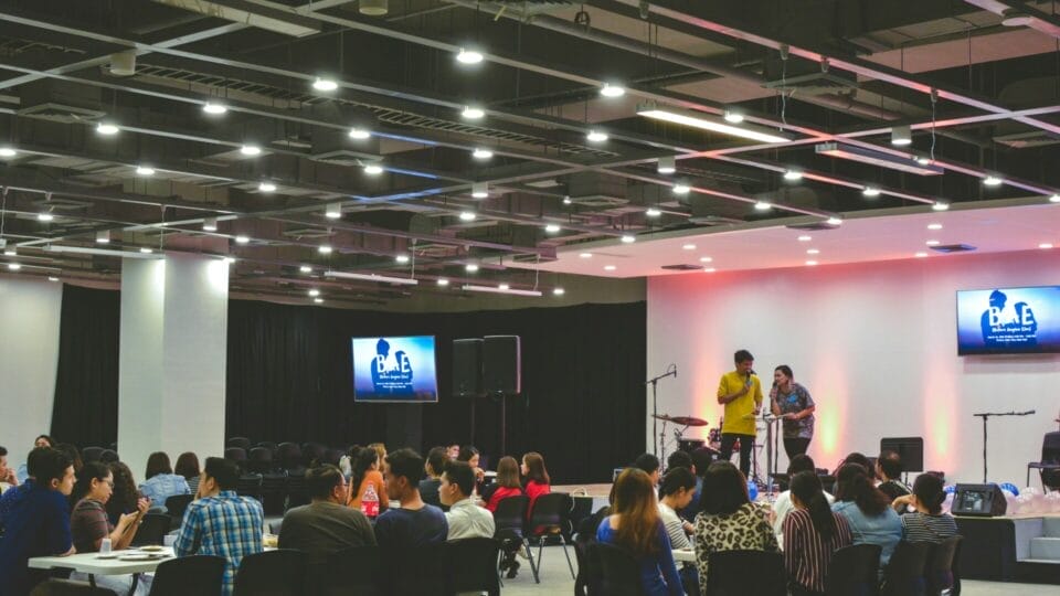 People sitting at tables in a large conference room, listening to speakers on a brightly lit stage with a presentation screen behind them.