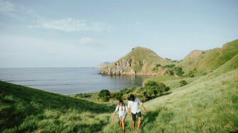 Two people hike up a grassy hill near the ocean with rocky cliffs and blue sky in the background.